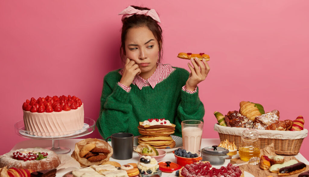 Unhappy ethnic girl looks with temptation at delicious dessert, sad as can put on weight after eating high caloried food, has variety confectionery and pasrty on table, poses in room with pink walls